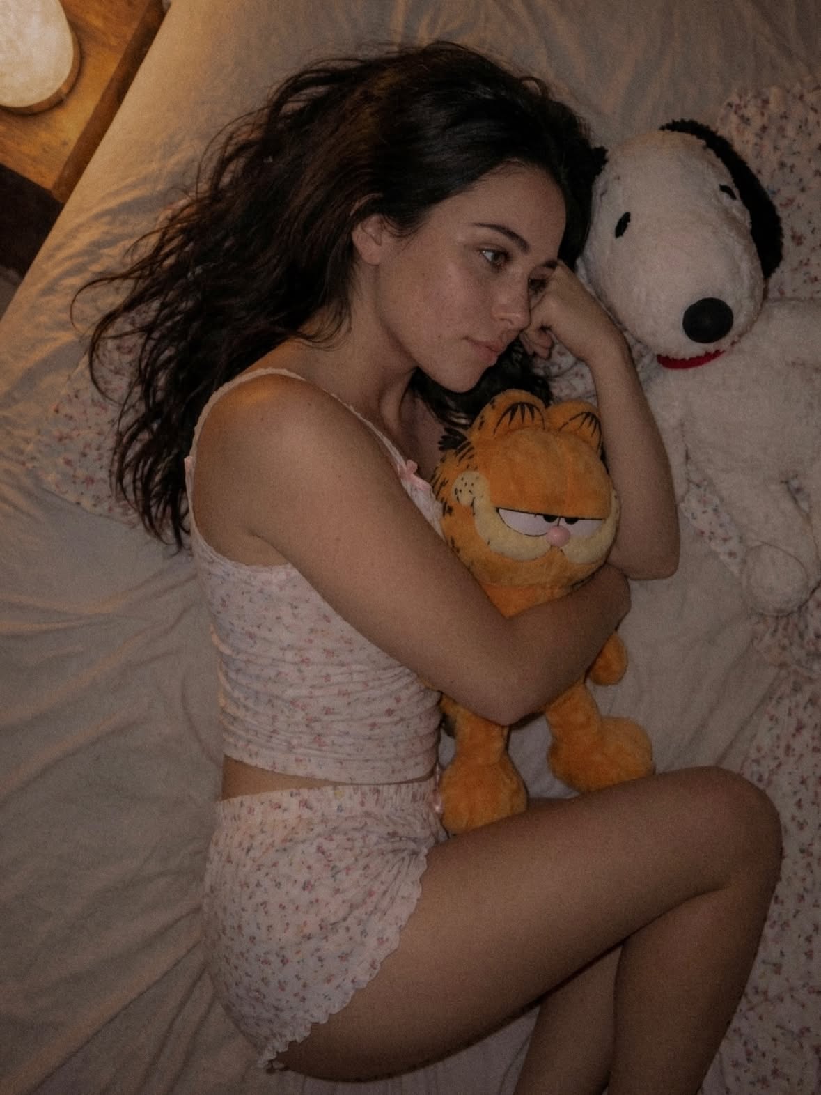 An intimate portrait series of a young woman with natural freckles, dark tousled hair, and light eyes, lying comfortably on a beige bed with floral bedding. She wears a delicate pink floral top and matching sleep shorts. She looks directly into the camera with a nostalgic, relaxed expression while tightly hugging a large Garfield plush toy, with a white Snoopy plush leaning on her arm. The atmosphere is warm, cozy, and personal, shot with shallow depth of field, natural evening indoor lighting, and a grainy, analogue film photography aesthetic.