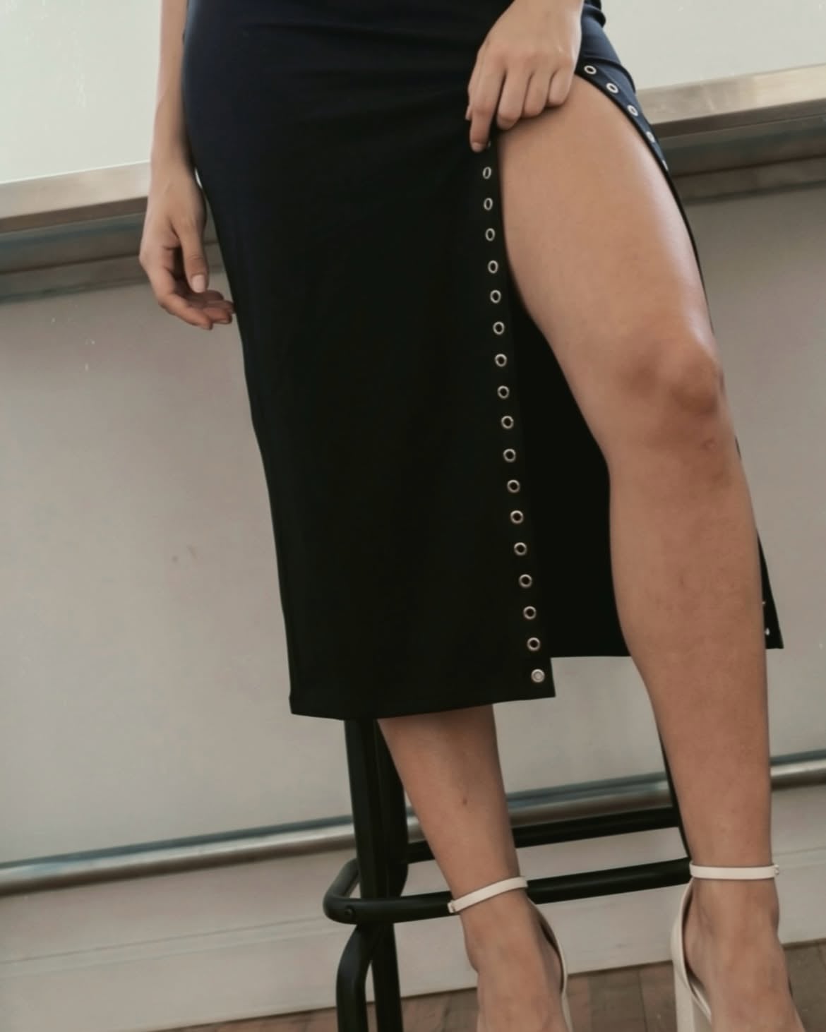 A young woman with long, wavy dark hair posing in a bright, minimalist studio. She is wearing a trendy black cutout crop top and a matching black midi skirt featuring a high leg slit with silver grommet details. She completes the look with statement white platform sandals. The setting includes a vintage oriental rug, wooden floors, and industrial-style furniture, creating a high-fashion, edgy aesthetic
